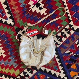 May include: A beige leather shoulder bag with a gold buckle and a white tag that says "Rivage" on it. The bag is on a colorful rug.