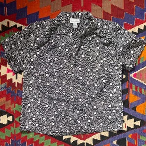 May include: A black and white polka dot button-up shirt with short sleeves. The shirt has a collar and a button closure. The brand name "Pierre Cardin" is visible on the label.