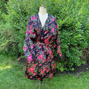 May include: A black satin robe with a floral pattern of pink and red roses, purple accents, and green leaves. The robe has a black lace trim on the collar and cuffs, and a matching belt. The robe is displayed on a vintage dress form.