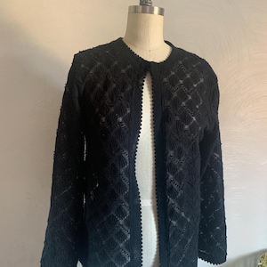 Puede incluir: Chaqueta negra abierta con un delicado patrón calado. La chaqueta presenta mangas largas y un ribete festoneado en los bordes. Se muestra en un maniquí, mostrando su ajuste a medida y su intrincado diseño. Es una prenda de vestir clásica.