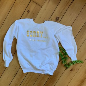 May include: White sweatshirt with long sleeves. The text "SORRY... I'd rather masturbate." is printed in gold foil across the chest. The sweatshirt is lying on a wooden surface.
