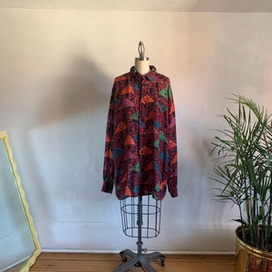 May include: A long-sleeved button-down shirt with a geometric pattern in shades of red, orange, purple, and blue. The shirt is on a mannequin and has a relaxed fit.