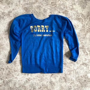 May include: A blue sweatshirt with the text "SORRY... I'd rather masturbate" in gold lettering. The long-sleeved top is made of a soft material and is laid flat on a patterned surface.