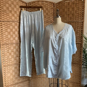 May include: A light blue pajama set, featuring a button-down top and matching pants, displayed on a mannequin and a hanger. The set has a floral pattern and is made of a silky material. The background includes a woven room divider and a houseplant.