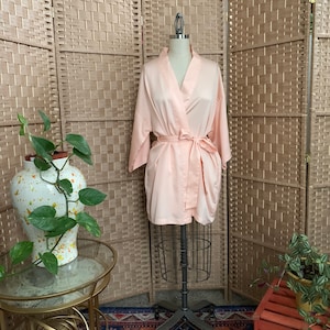 May include: A light peach-colored satin robe displayed on a vintage dress form. The robe has a matching tie belt and is surrounded by a woven room divider, a decorative table, and a patterned rug.