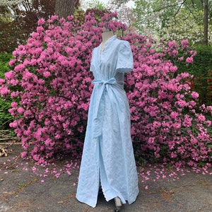 May include: A light blue, long,  sleeveless,  wrap dress with a white lace trim. The dress is tied at the waist with a matching sash. The dress is photographed in front of a bush of pink flowers.