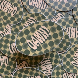 May include: A close-up of several oval-shaped stickers with a green and brown polka dot pattern. The stickers have the word "DAMN!" written in a stylized font.