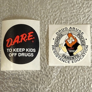 May include: Two round stickers with a black and white color scheme. The first sticker has the text "DA.RE." in red with the words "TO KEEP KIDS OFF DRUGS" below. The second sticker has a cartoon lion with the text "DRUG ABUSE SCHOOL POLICE RESISTANCE PARENTS EDUCATION" around the outside.