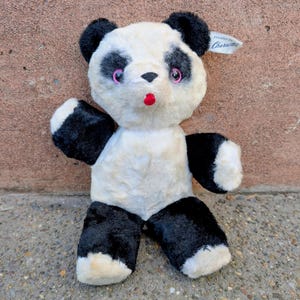 May include: A vintage panda bear plush toy with black and white fur. The panda has black ears, eye patches, and limbs, with a white body. It has pink eyes and a red mouth. A tag is attached to the ear.