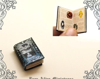 May include: A miniature book titled "Magical Stones & Their Properties" with a black cover and blue and silver accents. The book is open to a page with illustrations of different gemstones.