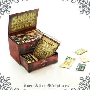 Puede incluir: Un cofre de madera en miniatura con cajones, lleno de art&iacute;culos en miniatura, incluyendo un libro titulado "A Treasury of Embroidery". El cofre est&aacute; decorado con un acabado de pintura desgastada y tiene una manija de metal. Tambi&eacute;n hay varios trozos peque&ntilde;os de papel en la mesa.