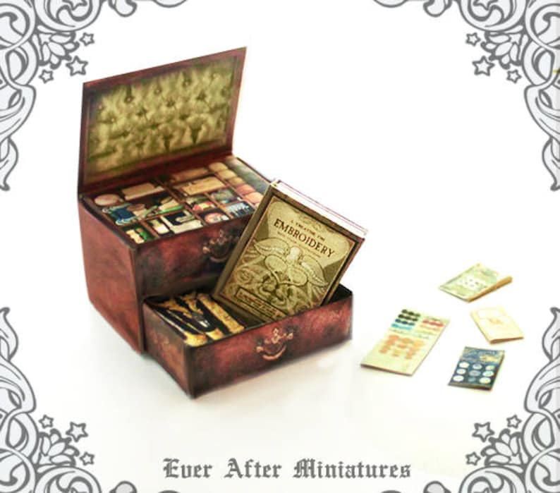 Puede incluir: Un cofre de madera en miniatura con cajones, lleno de art&iacute;culos en miniatura, incluyendo un libro titulado "A Treasury of Embroidery". El cofre est&aacute; decorado con un acabado de pintura desgastada y tiene una manija de metal. Tambi&eacute;n hay varios trozos peque&ntilde;os de papel en la mesa.