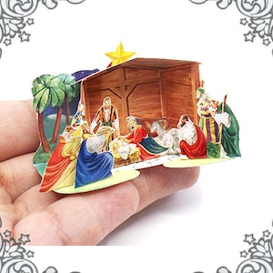 May include: A paper pop-up nativity scene with a wooden stable, a manger with a baby Jesus, and figures of Mary, Joseph, and the three wise men. The scene is set against a backdrop of palm trees and a starry night sky.