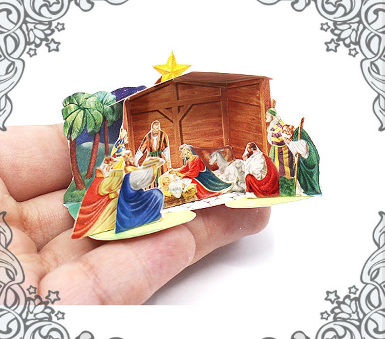 May include: A paper pop-up nativity scene with a wooden stable, a manger with a baby Jesus, and figures of Mary, Joseph, and the three wise men. The scene is set against a backdrop of palm trees and a starry night sky.