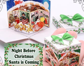 Può includere: Un biglietto pop-up con una scena natalizia con Babbo Natale e le renne. Il biglietto è in una scatola bianca con un fiocco verde e decorazioni di agrifoglio. Il testo sul biglietto recita "Night Before Christmas Santa is Coming".