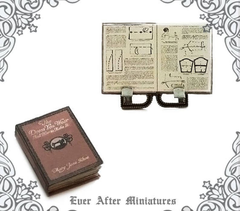 May include: A miniature book titled "The Dress You Wear And How to Make It" by Mary Jane Rhoe. The book is closed and has a brown cover with a sewing machine illustration. A miniature open book with sewing instructions is also pictured.