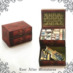 Puede incluir: Una caja de madera con dos cajones y una tapa con bisagras. La caja est&aacute; abierta y el interior est&aacute; lleno de peque&ntilde;os compartimentos y art&iacute;culos. La caja es marr&oacute;n con detalles dorados. El texto "Ever After Miniatures" est&aacute; impreso en la parte inferior de la imagen.