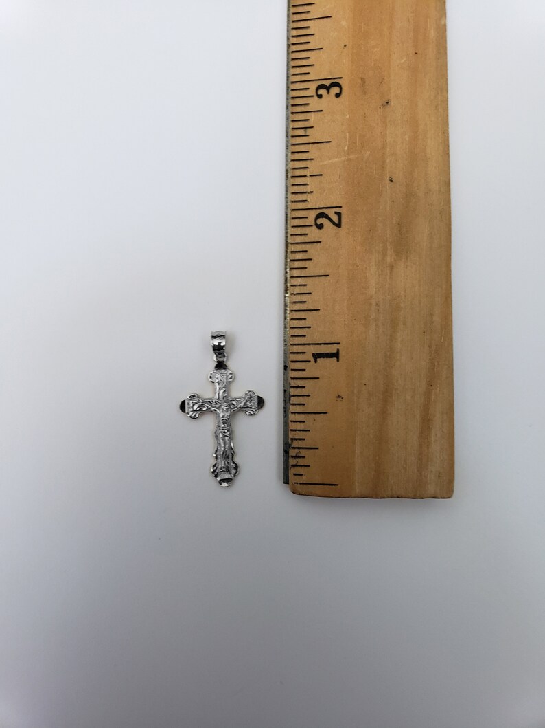 May include: A silver cross pendant with a detailed design, measuring approximately 2.5 cm in length. The cross is shown next to a ruler for size reference.