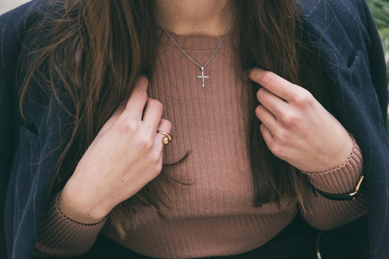 Pode incluir: Uma mulher vestindo um su&eacute;ter marrom e um blazer azul marinho com um colar de cruz de prata e um anel de ouro no dedo.