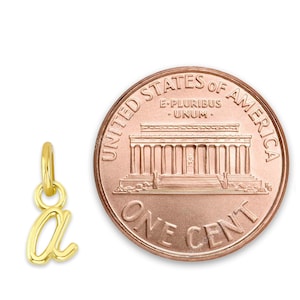 May include: A small, gold-coloured charm in the shape of the lowercase letter "a" hangs next to a U.S. one-cent coin. The charm is likely made of gold and has a polished finish. The coin is copper-coloured and shows the Lincoln Memorial.