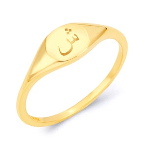 May include: A gold signet ring featuring an Arabic letter engraved on the top. The ring is oval shaped and has a smooth, polished finish.