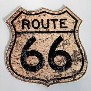 Könnte beinhalten: Ein braunes und schwarzes Route 66 Patch mit einem abgenutzten Look. Das Patch ist schildförmig und hat einen weißen Hintergrund mit schwarzem Text und Zahlen.