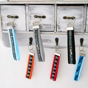 May include: Six colorful keychains with different designs hanging on hooks. The keychains are made of fabric with various patterns, including skulls, cameras, and stars. The keychains are attached to silver key rings.