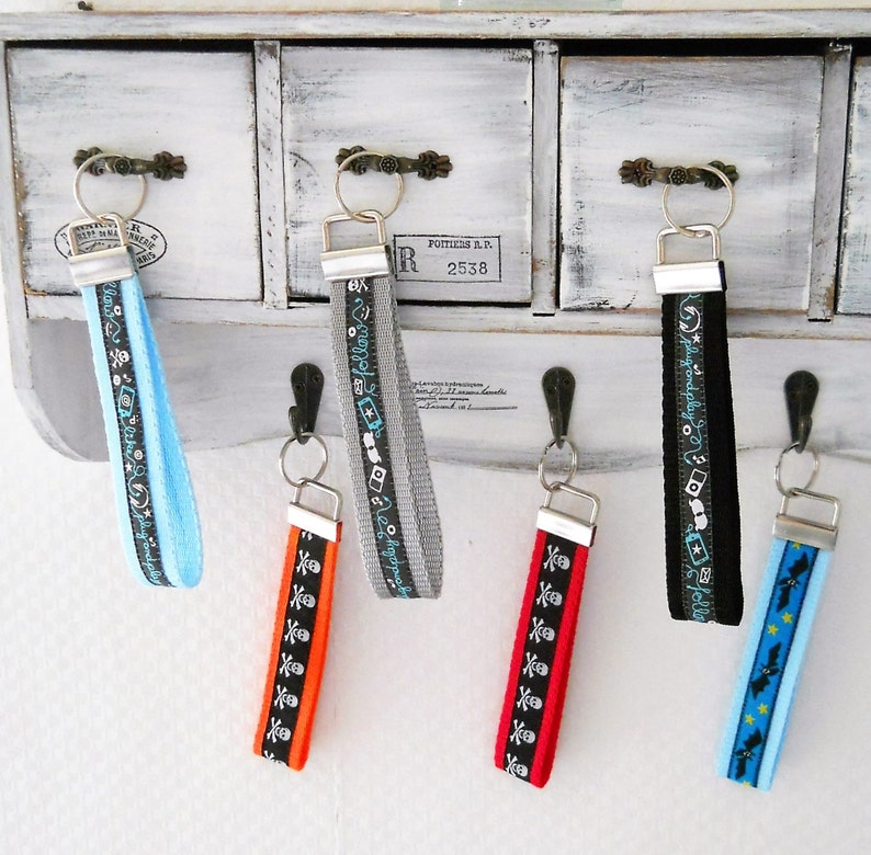 May include: Six colorful keychains with different designs hanging on hooks. The keychains are made of fabric with various patterns, including skulls, cameras, and stars. The keychains are attached to silver key rings.