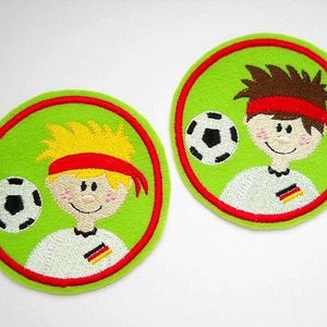 May include: Two green felt circles with red borders, each featuring a cartoon football player with a football. One player has blonde hair and the other has brown hair. Both players are wearing red headbands and white jerseys with the German flag.