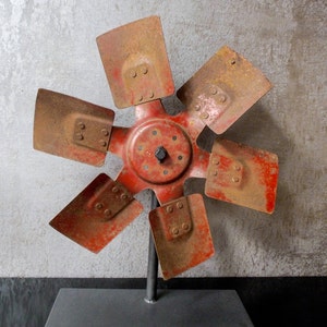 Könnte beinhalten: Eine rostige rote Metall-Lüfterblatt-Skulptur mit sechs Blättern, die auf einem schwarzen Metallständer montiert sind. Die Lüfterblätter sind in einem Sternburst-Muster angeordnet.