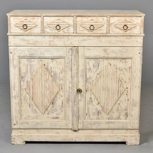 May include: A distressed, off-white cabinet with four small drawers above two doors. The drawers have ring pulls and diamond-shaped carvings. The doors feature diamond-shaped panels. The cabinet has a vintage, rustic aesthetic.