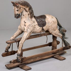 May include: A vintage rocking horse with a distressed, speckled white and brown finish. The horse features a dark saddle and a flowing, gray-green mane and tail. The wooden base is weathered, adding to its antique charm.