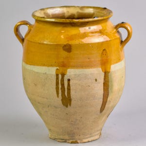 以下が含まれることがあります： 2つの取っ手が付いた、大きなアンティークの陶器の壺。壺は主にクリーム色で、上部に幅広の黄色い帯と黄色の塗料の滴があります。縁と取っ手も黄色です。壺は素朴で古びた外観をしています。