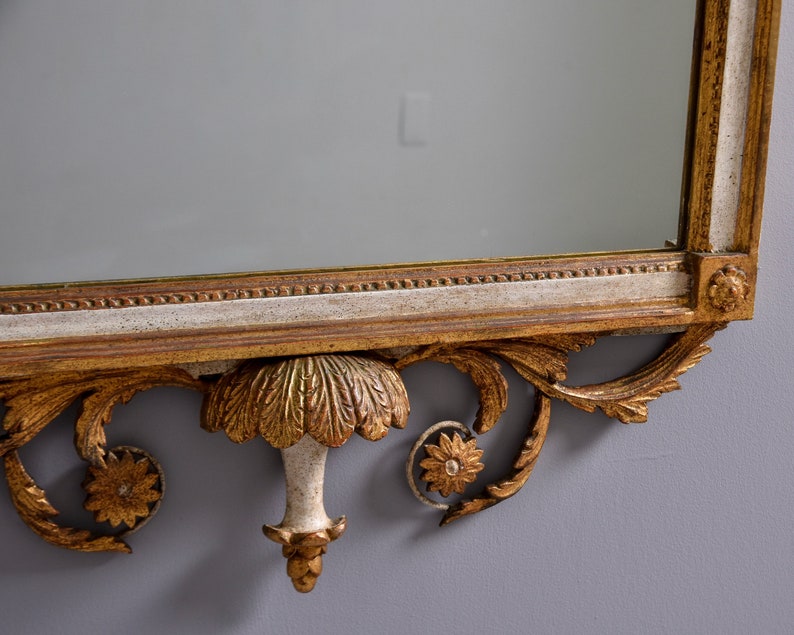 May include: A gold and white ornate mirror with a decorative carved frame. The frame features a central floral design and scrolling details.