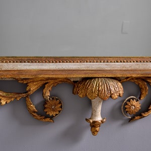 May include: A gold-painted wooden mirror with an ornate, carved frame. The frame features a central, leaf-shaped design with a decorative finial.