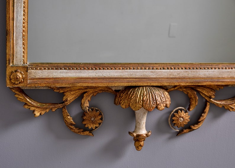 May include: A gold-painted wooden mirror with an ornate, carved frame. The frame features a central, leaf-shaped design with a decorative finial.