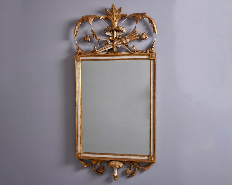 May include: A gold and white ornate mirror with a decorative top featuring a floral design and a central motif of a stylized fan. The mirror has a rectangular frame with a beveled edge.