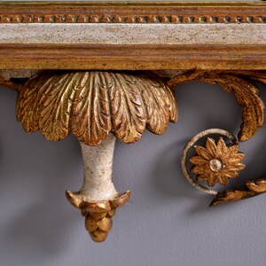 May include: Close-up of a decorative, ornate, gold-painted wooden shelf bracket with a white painted center. The bracket features a carved, leaf-like design and a central, stylized flower.