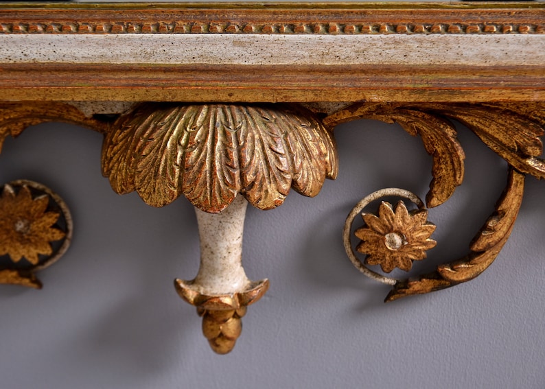 May include: Close-up of a decorative, ornate, gold-painted wooden shelf bracket with a white painted center. The bracket features a carved, leaf-like design and a central, stylized flower.