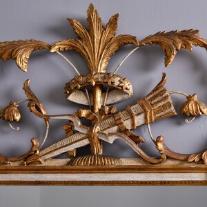 May include: A gold and white ornate mirror with a carved wooden frame. The frame features a central design of a stylized arrow and a crown with floral accents.