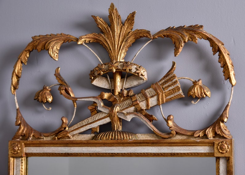 May include: A gold and white ornate mirror with a carved wooden frame. The frame features a central design of a stylized arrow and a crown with floral accents.