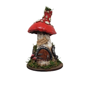 Puede incluir: Una casa de gnomo de cerámica con forma de seta roja y blanca con una base de madera marrón. La casa tiene una pequeña puerta con marco de metal y un letrero que dice "Gnome house".