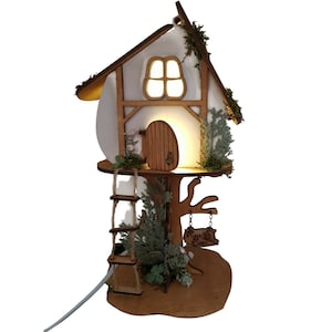 May include: A wooden fairy house with a white roof and a light inside. The house is on a tree trunk with a ladder and a swing. There are green leaves and branches around the house.