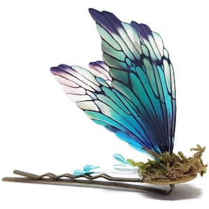 Puede incluir: Horquilla decorativa con diseño de ala de mariposa. El ala presenta un degradado de azul, turquesa y morado, con delicados detalles de venas. La horquilla tiene una base color bronce con musgo y pequeños detalles azules.