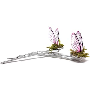 Puede incluir: Dos pinzas para el pelo plateadas con alas de mariposa rosa y blanca y detalles de musgo verde.