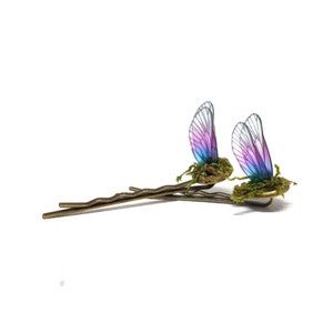 Puede incluir: Dos pinzas para el pelo de mariposa iridiscentes con alas rosas, azules y moradas. Las alas están unidas a un pequeño trozo de musgo y una horquilla dorada.