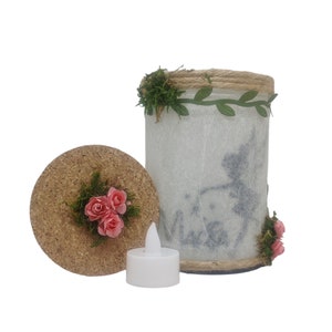 Puede incluir: Un portavelas cil&iacute;ndrico blanco con tapa de corcho y dise&ntilde;o floral. El portavelas est&aacute; decorado con un ribete de cuerda, hojas verdes y flores rosas. Se incluye una peque&ntilde;a vela de t&eacute; blanca.