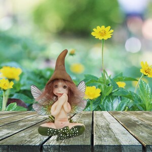 May include: A small, brown-haired fairy figurine with wings, wearing a brown pointed hat and a green and white polka dot dress. The fairy is sitting with its hands clasped in front of its chest. The figurine is on a wooden surface with a blurred background of green foliage and yellow flowers.