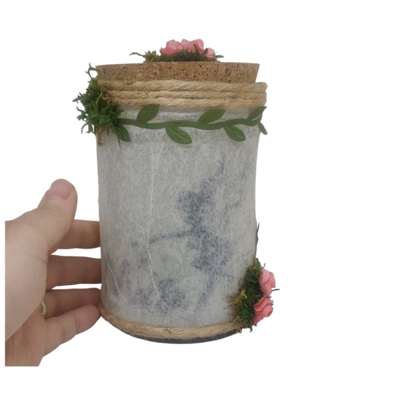 Puede incluir: Un tarro blanco con tapa de corcho decorado con flores rosas, hojas verdes y cuerda. El tarro tiene un estilo r&uacute;stico y de granja.