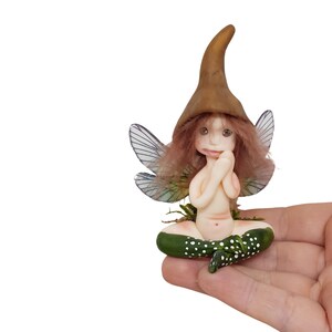 May include: A small fairy figurine with brown hair, wearing a brown pointed hat and sitting cross-legged. The fairy has wings with a green and blue iridescent pattern and is holding its hands together in front of its face.